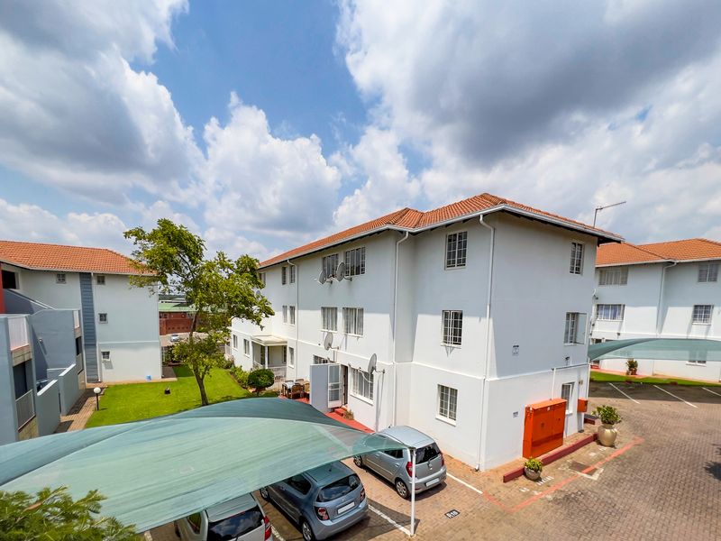 Exterior view of a multi-story apartment complex featuring a tiled roof, paved parking area with shade cloth carports, and a communal garden space.