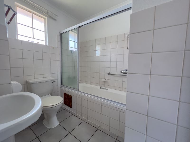 A functional bathroom featuring a pedestal basin, toilet, and a bathtub with a sliding glass screen, finished with white ceramic wall and floor tiles.
