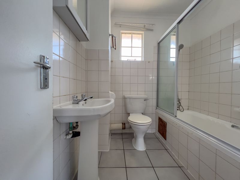 A functional bathroom featuring a pedestal basin, toilet, and a bathtub with a shower screen, finished with white wall tiles and floor tiles.