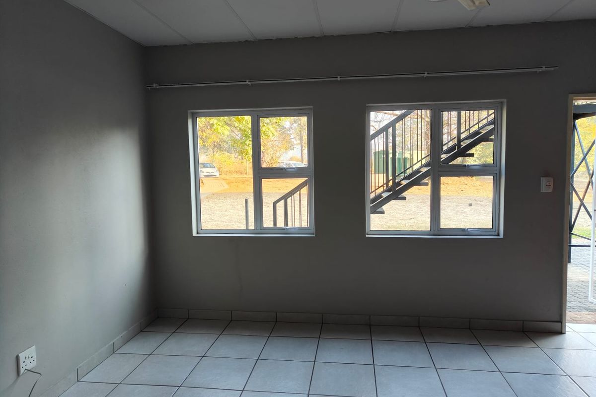 An empty living area featuring neutral tiled flooring, grey walls, and large windows providing natural light and views of the exterior staircase.