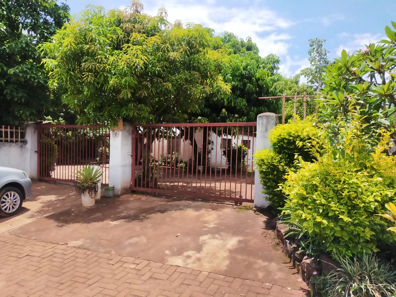 Front entrance view featuring a metal driveway gate, masonry pillars, and a paved driveway surrounded by established garden greenery.