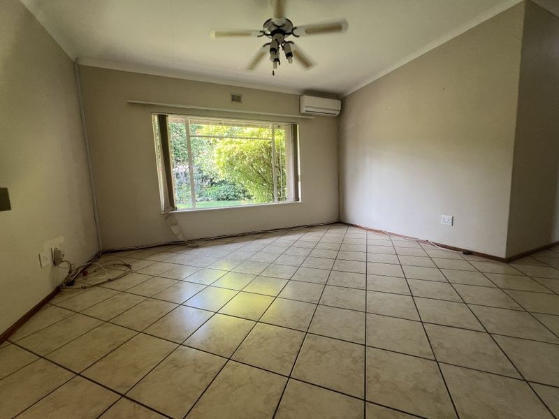 An empty living room featuring tiled flooring, a ceiling fan, and a wall-mounted air conditioning unit, with a large window overlooking the garden.
