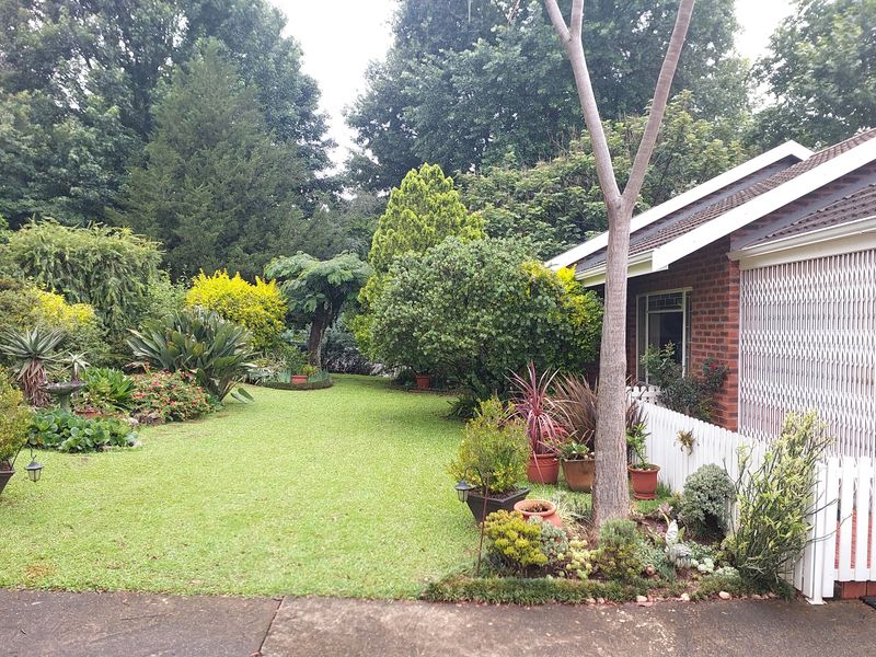 A well-maintained garden area featuring a lush lawn, mature trees, and a brick townhouse exterior with a white picket fence.