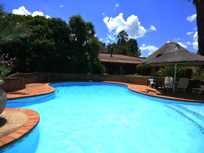 Outdoor swimming pool area featuring a brick-paved surround, a thatched lapa, and established garden landscaping.