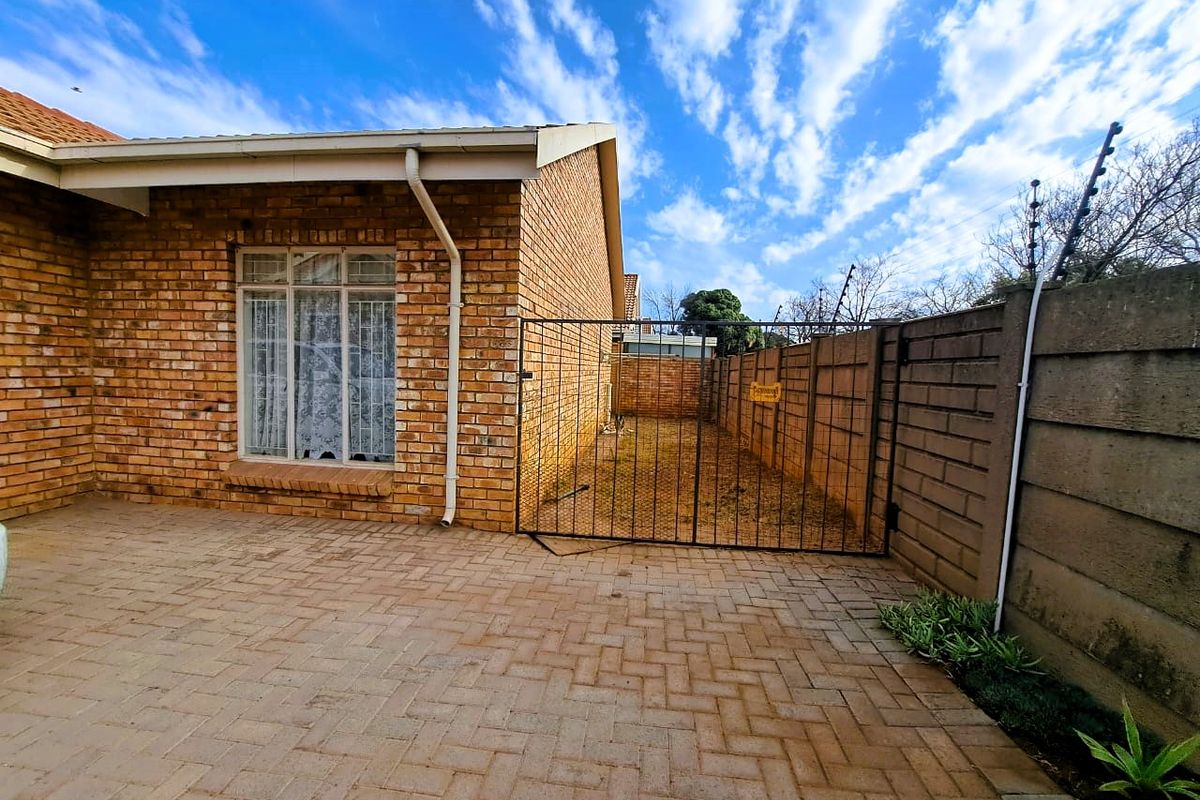 A rear exterior view of the townhouse featuring a paved patio area, facebrick facade, and a secure gated side yard with perimeter electric fencing.