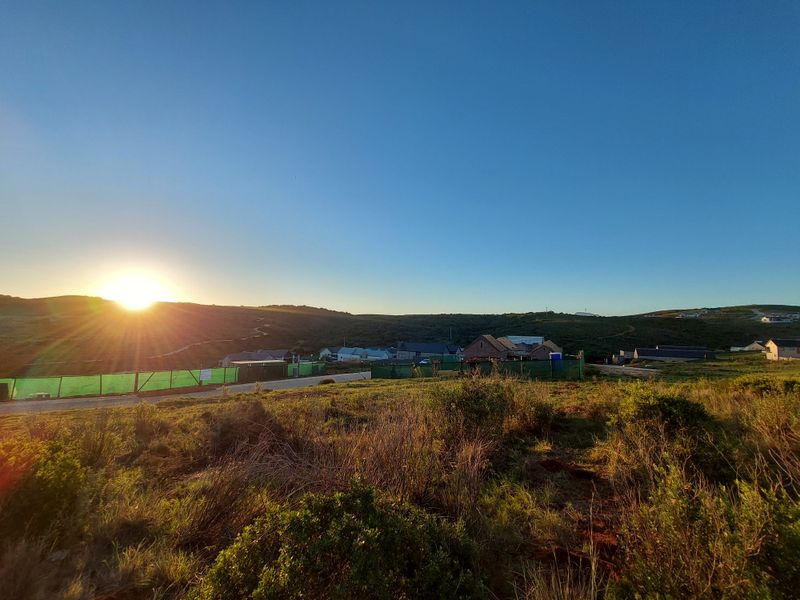 A scenic landscape view of a coastal or rural residential area at sunrise, featuring rolling hills and distant housing developments.