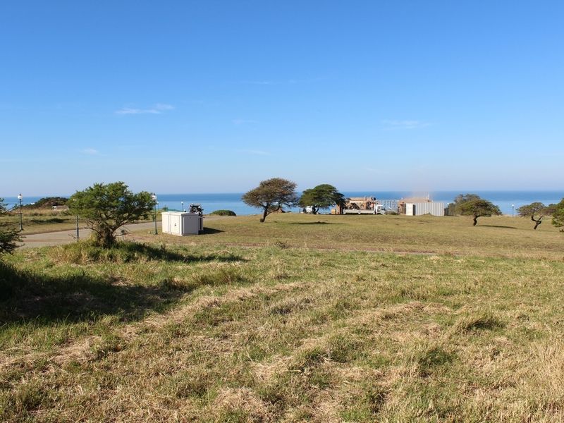 A wide-angle view of a vacant residential plot featuring natural grass terrain, scattered trees, and a scenic ocean horizon in the background.