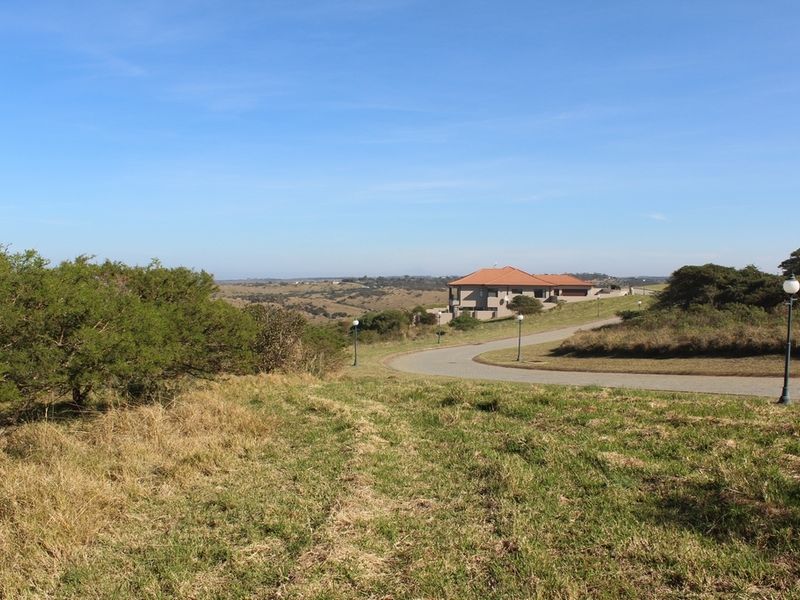 A scenic view of a vacant residential plot featuring a paved driveway, street lighting, and surrounding natural landscape with distant housing.