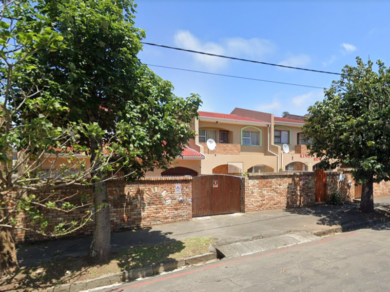 Front exterior view of a townhouse complex featuring a brick boundary wall, wooden entrance gate, and tiled roofing.