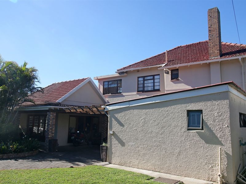Rear view of the property featuring a tiled roof, brick chimney, paved driveway, and a covered pergola area with established garden surroundings.