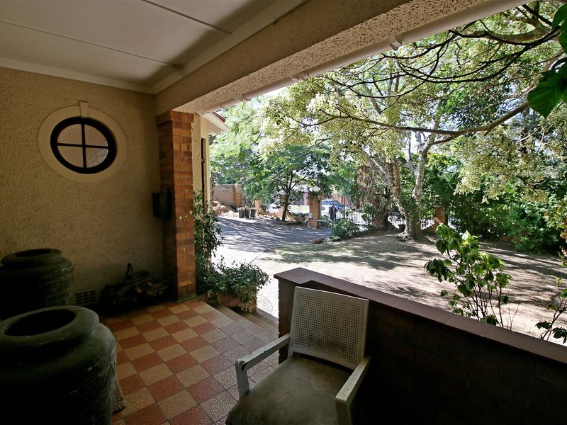 A covered patio area featuring brick pillars, a unique porthole window, and tiled flooring, overlooking a shaded front garden and driveway.