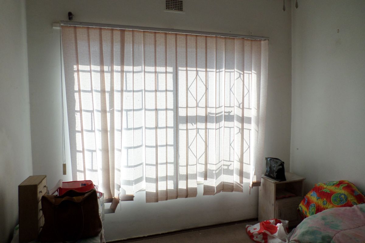 A bedroom featuring a window with security bars and vertical blinds, requiring cosmetic updates to improve the overall aesthetic and condition of the space.