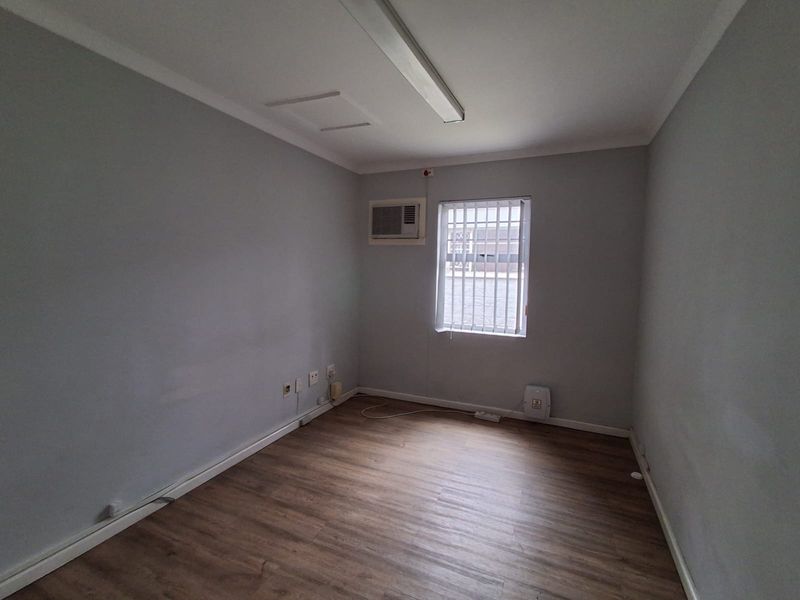 A clean, empty office space featuring laminate flooring, an integrated air conditioning unit, and a window with security bars.