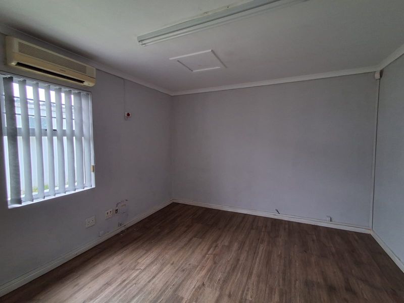 A vacant office space featuring laminate flooring, a wall-mounted air conditioning unit, and vertical window blinds, providing a clean and functional workspace environment.