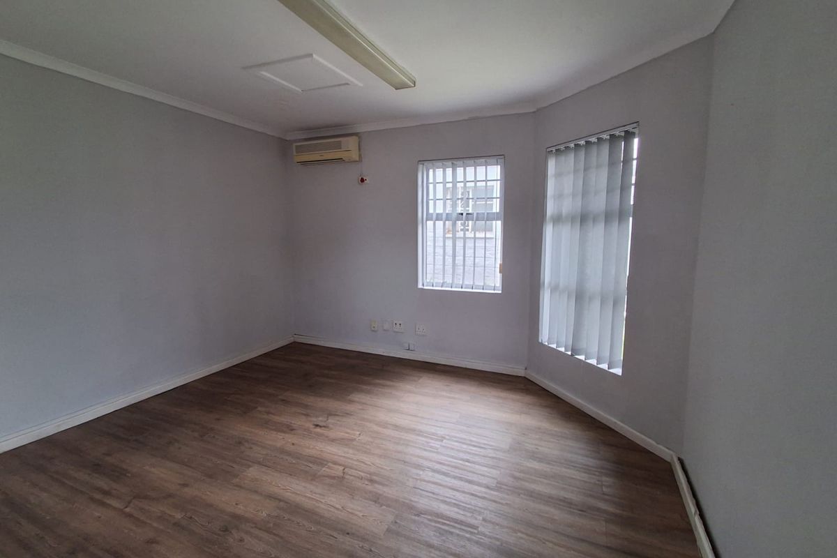 A clean, empty office space featuring wood-look laminate flooring, an air conditioning unit, and windows fitted with vertical blinds and security bars.
