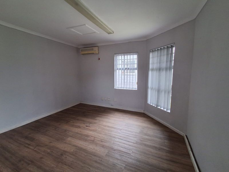 A clean, empty office space featuring wood-look laminate flooring, an air conditioning unit, and windows fitted with vertical blinds and security bars.