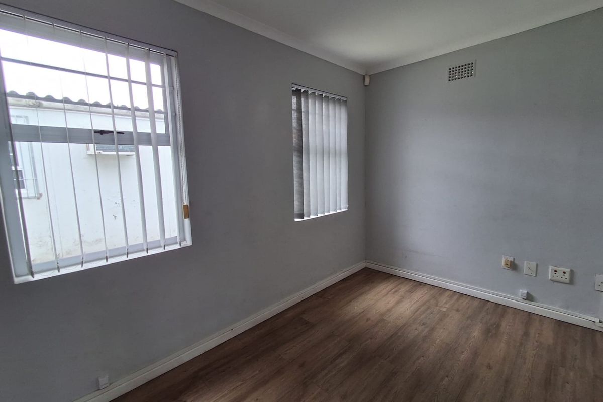 A clean, empty office space featuring wood-look laminate flooring, neutral walls, and windows fitted with vertical blinds and security bars.