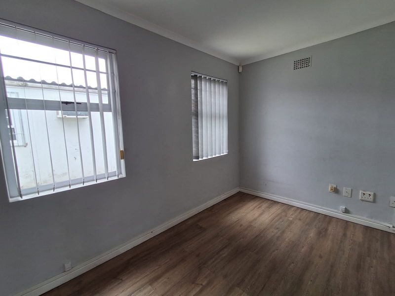 A clean, empty office space featuring wood-look laminate flooring, neutral walls, and windows fitted with vertical blinds and security bars.