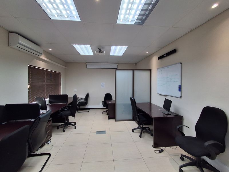 A functional office space featuring tiled flooring, suspended ceilings with recessed lighting, a wall-mounted air conditioning unit, and a built-in glass partition.