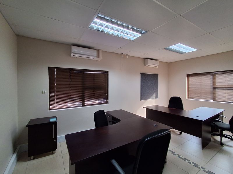 A functional office space featuring tiled flooring, suspended ceilings with recessed lighting, dual air conditioning units, and window blinds.