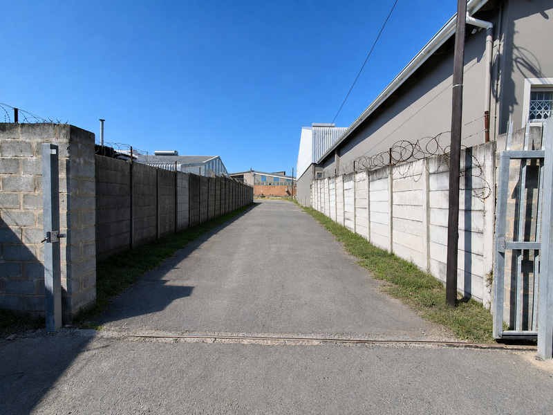 A paved industrial access driveway enclosed by high precast concrete boundary walls topped with security razor wire.