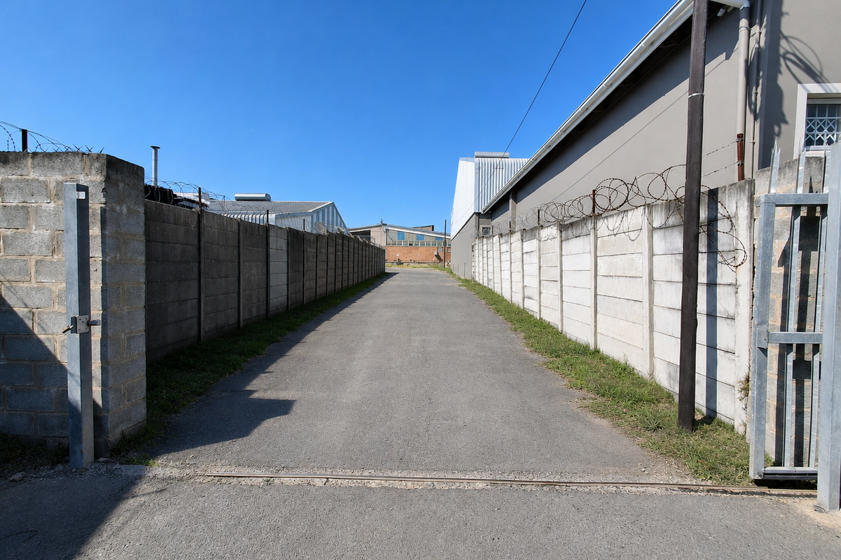 A paved industrial access driveway enclosed by high precast concrete boundary walls topped with security razor wire.