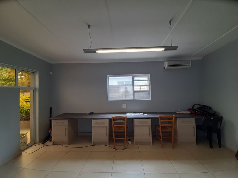 A functional office space featuring a long built-in desk with integrated cabinetry, tiled flooring, air conditioning, and ample natural light from the window and door.
