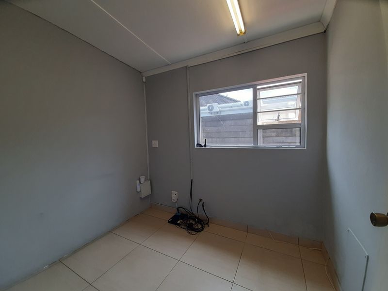 A compact, empty office space featuring tiled flooring, a fluorescent light fixture, and an aluminium window with security bars.