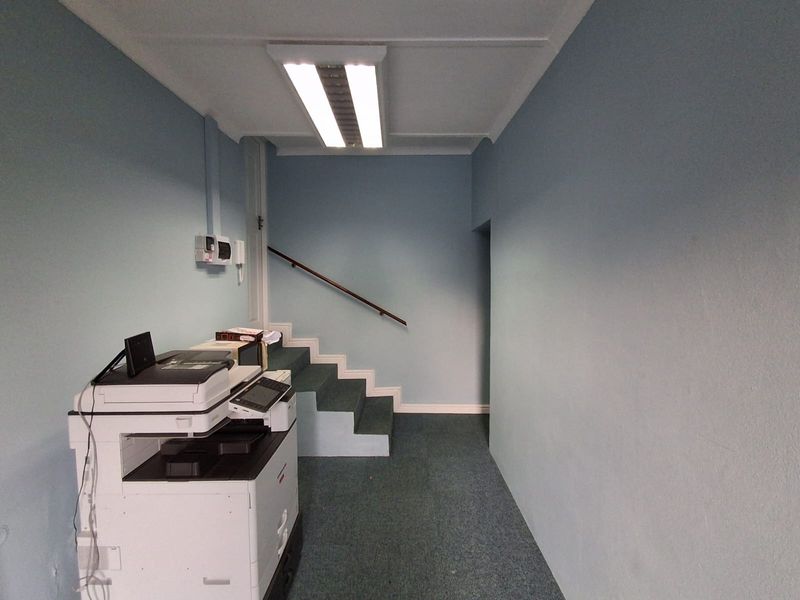 Office space featuring blue painted walls, carpeted flooring, and a staircase with a handrail, illuminated by overhead fluorescent lighting.