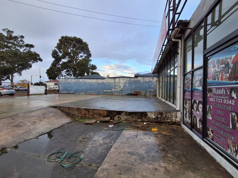 Exterior view of a retail property featuring a large glass storefront, concrete paved area, and a boundary wall requiring maintenance.