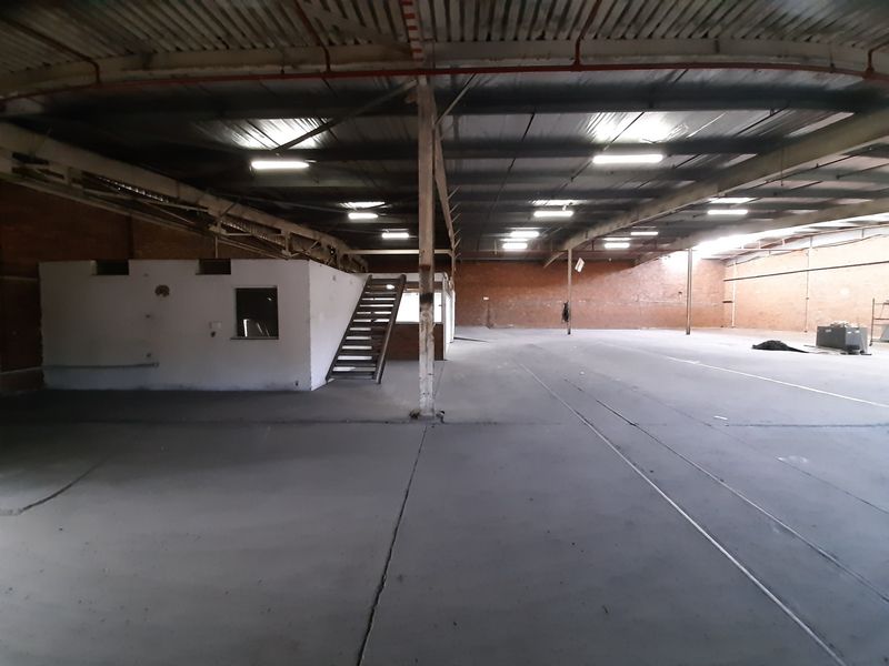 Expansive industrial warehouse space featuring high ceilings with exposed steel trusses, concrete flooring, and a mezzanine level with an integrated metal staircase.