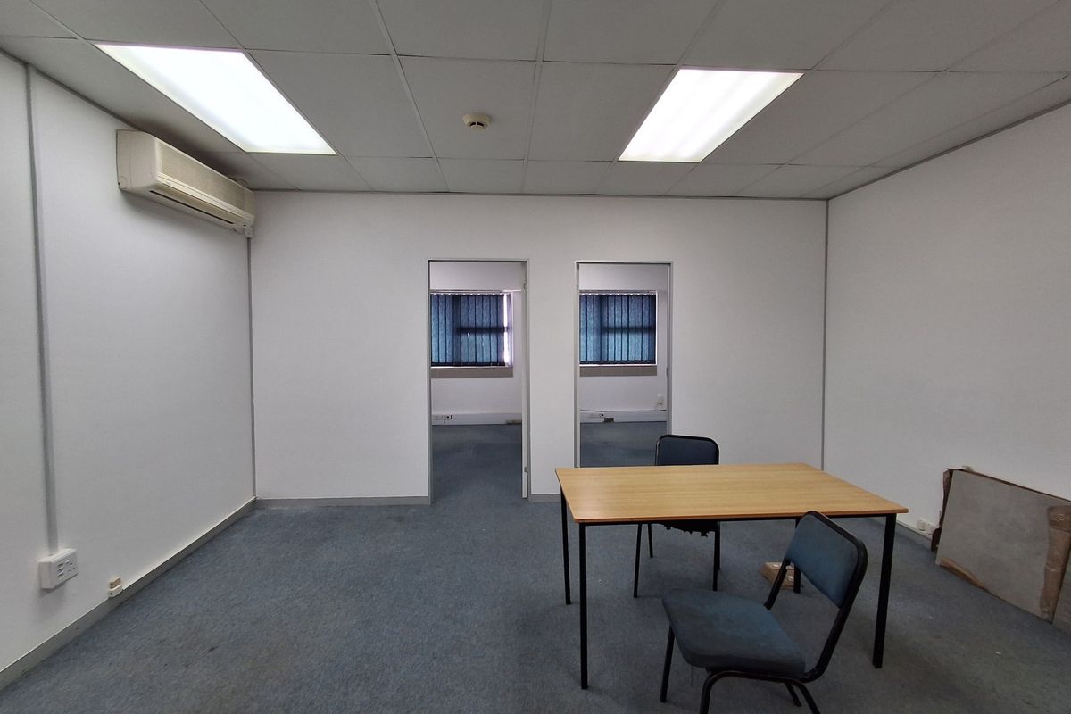 A functional, empty office space featuring a suspended ceiling, recessed lighting, wall-mounted air conditioning, and carpeted flooring with dual access points.