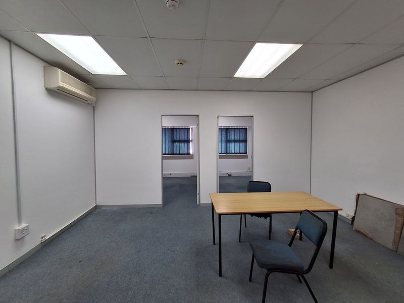A functional, empty office space featuring a suspended ceiling, recessed lighting, wall-mounted air conditioning, and carpeted flooring with dual access points.