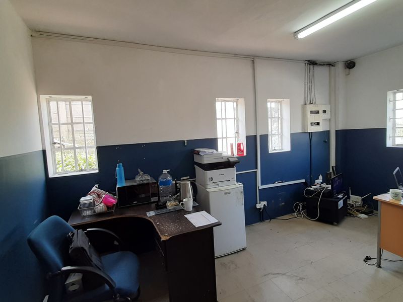 Functional office space featuring security bars on windows, exposed electrical distribution board, and basic fluorescent lighting, suitable for a business environment.