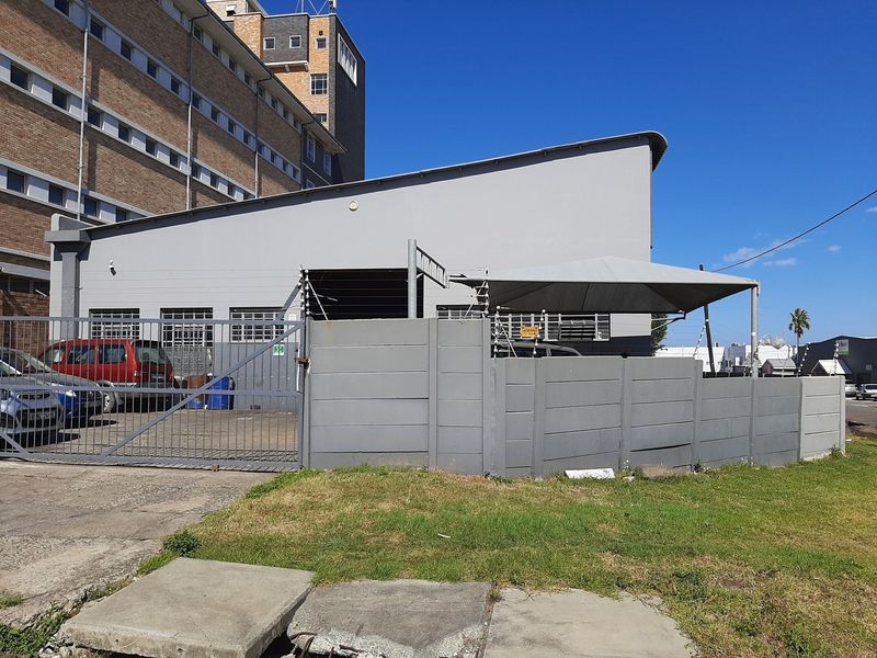 Rear exterior view of the commercial property featuring a secure gated entrance, concrete boundary wall with electric fencing, and a shaded parking area.