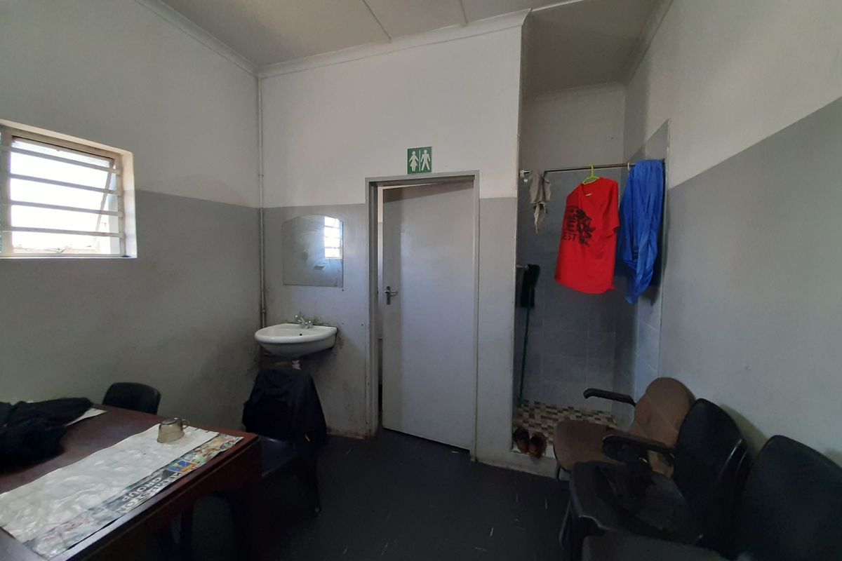 A functional commercial bathroom area featuring a wall-mounted basin, shower cubicle, and security-barred window, requiring cosmetic refurbishment and deep cleaning.