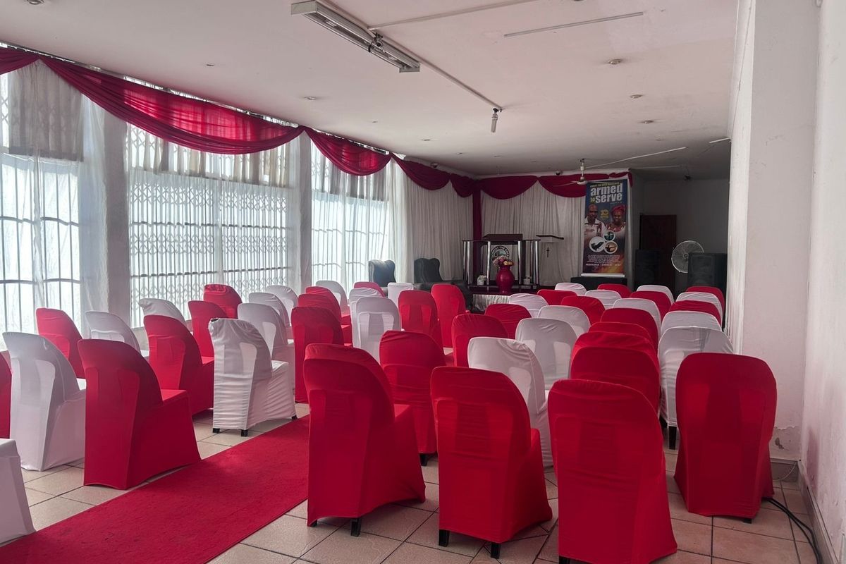 A spacious commercial hall featuring tiled flooring, recessed lighting, and large windows with security gates, currently set up for event seating.