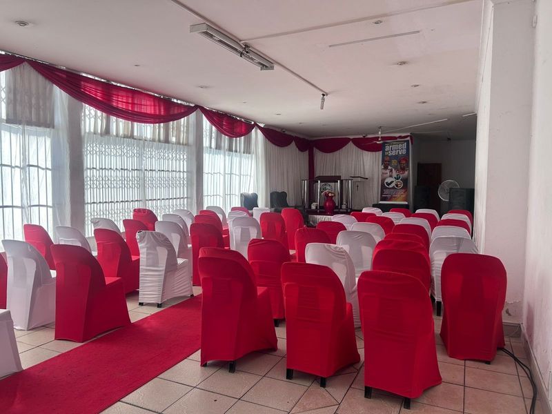 A spacious commercial hall featuring tiled flooring, recessed lighting, and large windows with security gates, currently set up for event seating.