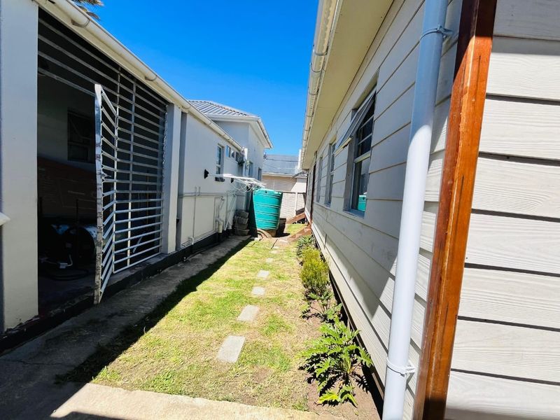 A narrow side passage between two building structures featuring weatherboard cladding, a water tank, and a small grassed area with paving stones.