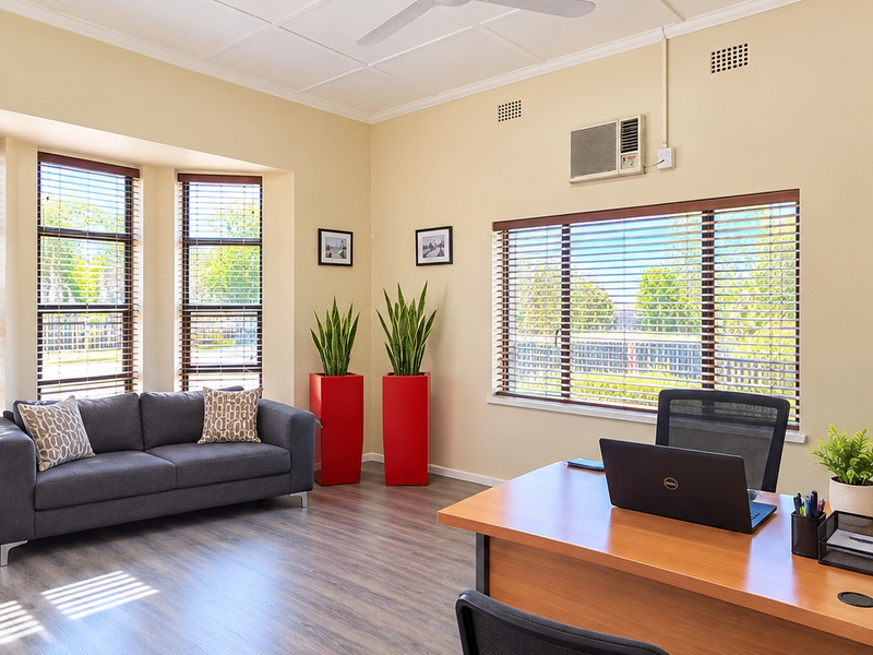 A bright and professional office space featuring laminate flooring, a bay window with venetian blinds, and integrated wall-mounted air conditioning.
