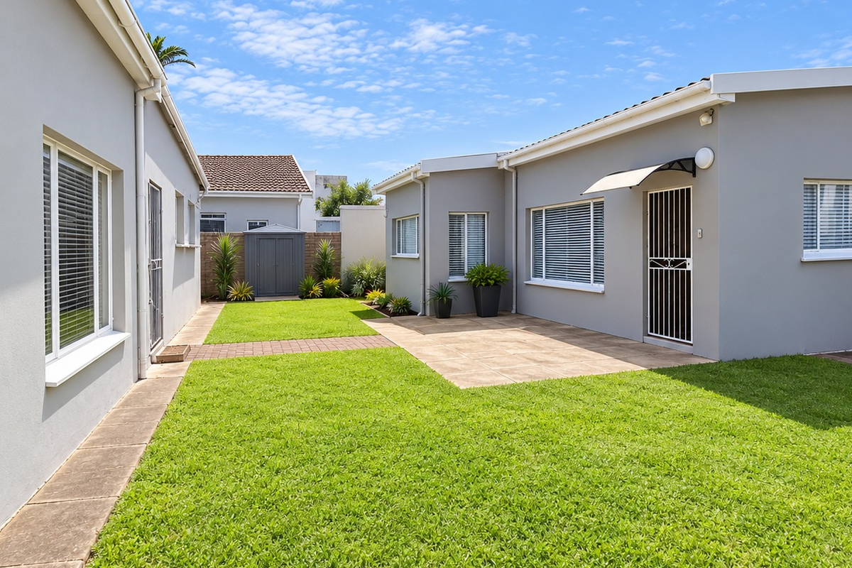 A well-maintained rear exterior view featuring a paved patio area, manicured lawn, and clean grey building facades with security gates and awnings.