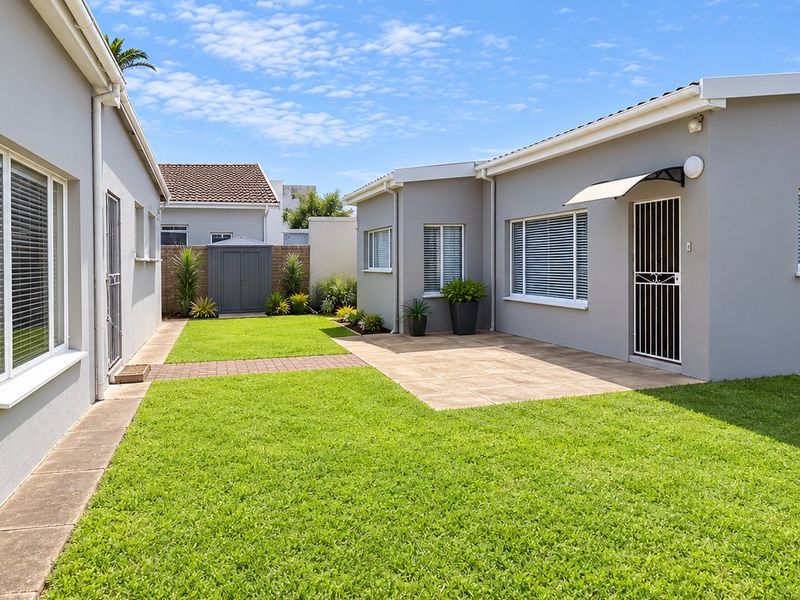 A well-maintained rear exterior view featuring a paved patio area, manicured lawn, and clean grey building facades with security gates and awnings.