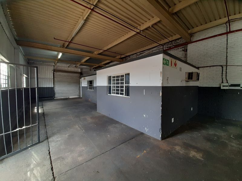Industrial warehouse space featuring high ceilings with exposed trusses, a secure roller shutter door, and a partitioned office area with security bars.