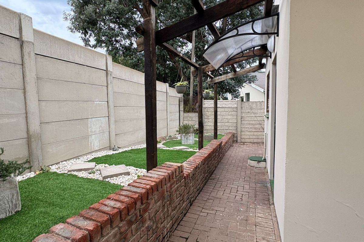 A rear exterior view of the cottage featuring a paved walkway, a brick retaining wall, and a timber pergola structure along the boundary wall.