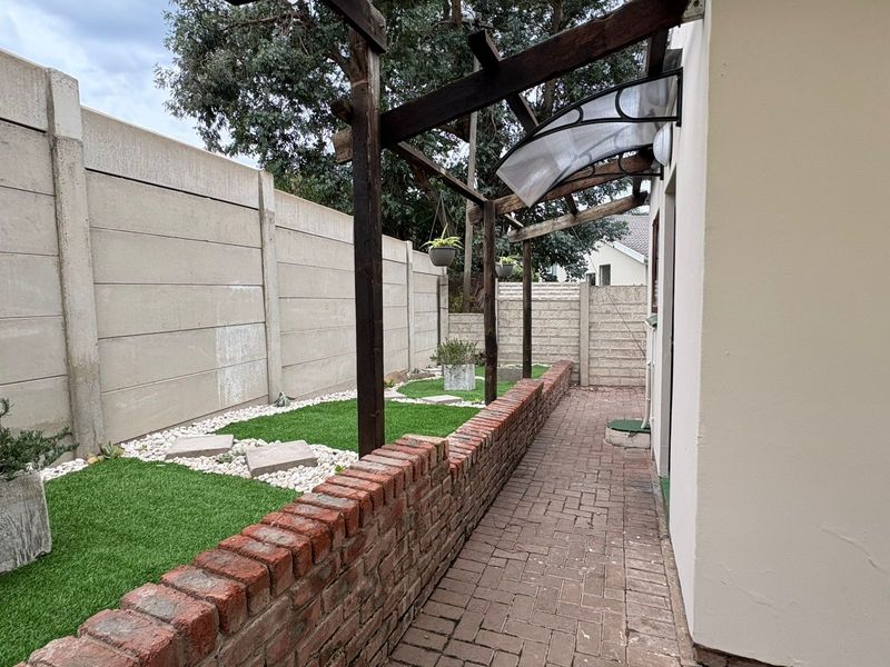 A rear exterior view of the cottage featuring a paved walkway, a brick retaining wall, and a timber pergola structure along the boundary wall.