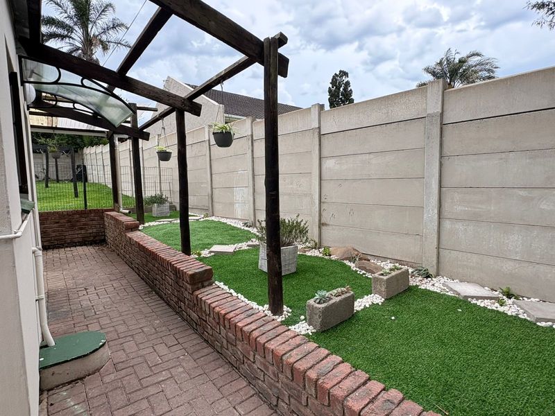 A private outdoor patio area featuring a timber pergola, brick retaining wall, and low-maintenance artificial lawn, enclosed by a high precast concrete boundary wall.