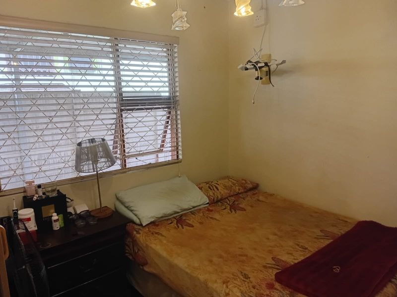 A compact bedroom featuring a window with security bars and venetian blinds, requiring minor cosmetic updates and electrical fixture maintenance.