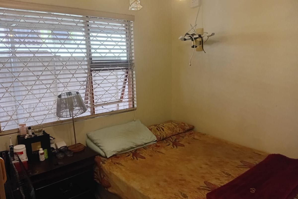 A compact bedroom featuring a window with security bars and venetian blinds, requiring minor cosmetic updates and electrical fixture maintenance.