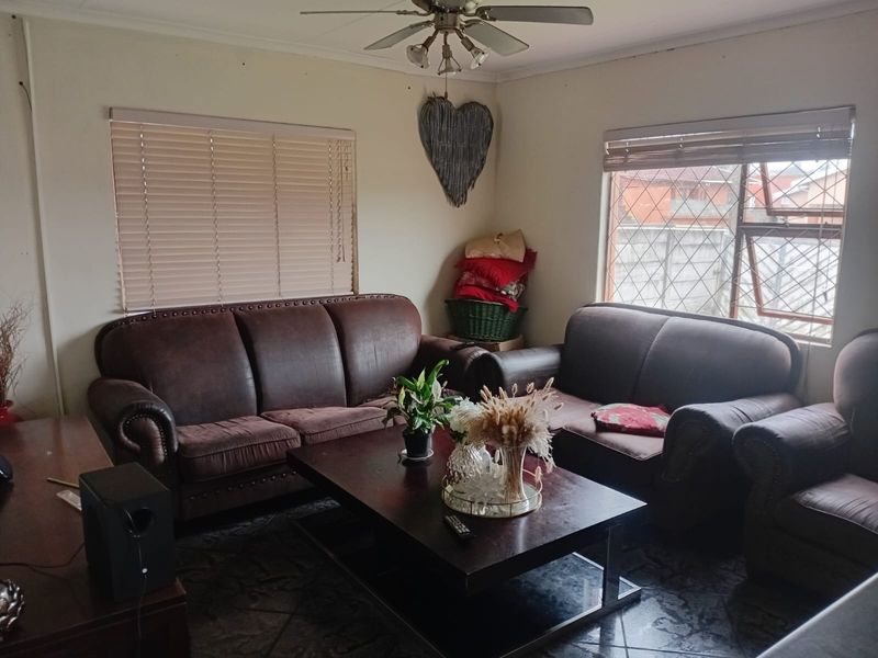 A comfortable living room featuring tiled flooring, a ceiling fan, and natural light provided by large windows with fitted venetian blinds.
