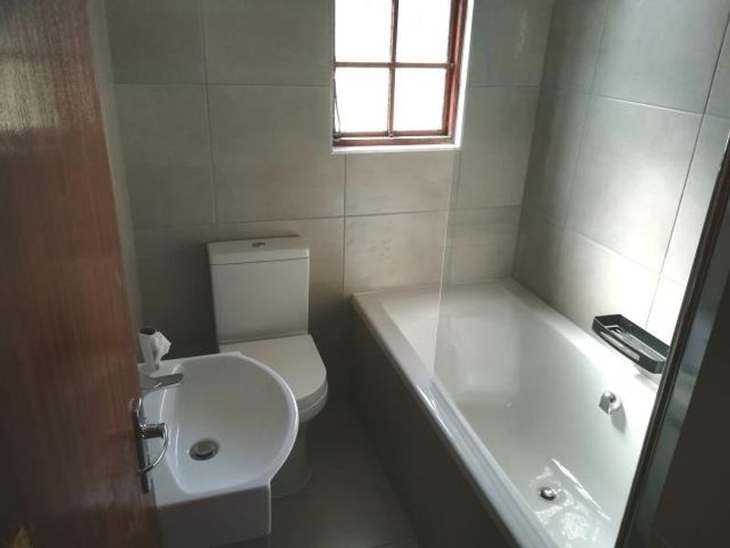 A clean, modern bathroom featuring a white bathtub with a glass screen, a pedestal washbasin, and a toilet, finished with neutral wall tiling.